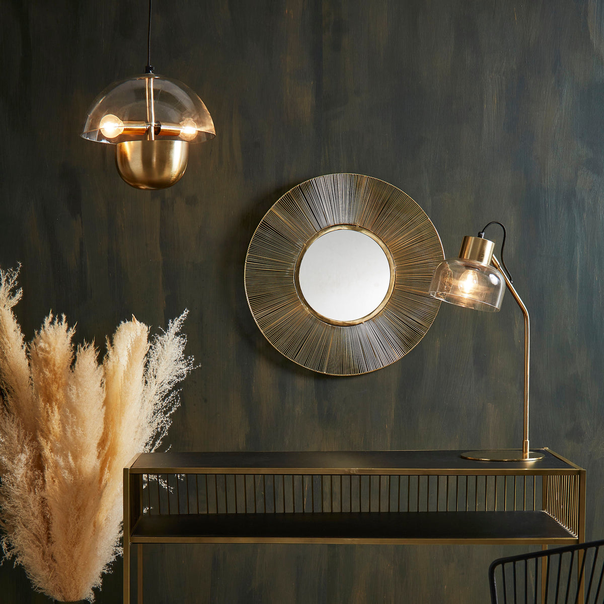 A styled interior scene featuring a brass console table against a dark textured wall, with a round sunburst wall mirror above it, a hanging glass pendant light, a floor lamp, a black wire chair with a patterned cushion, pampas grass in a vase, and a rug on the floor.