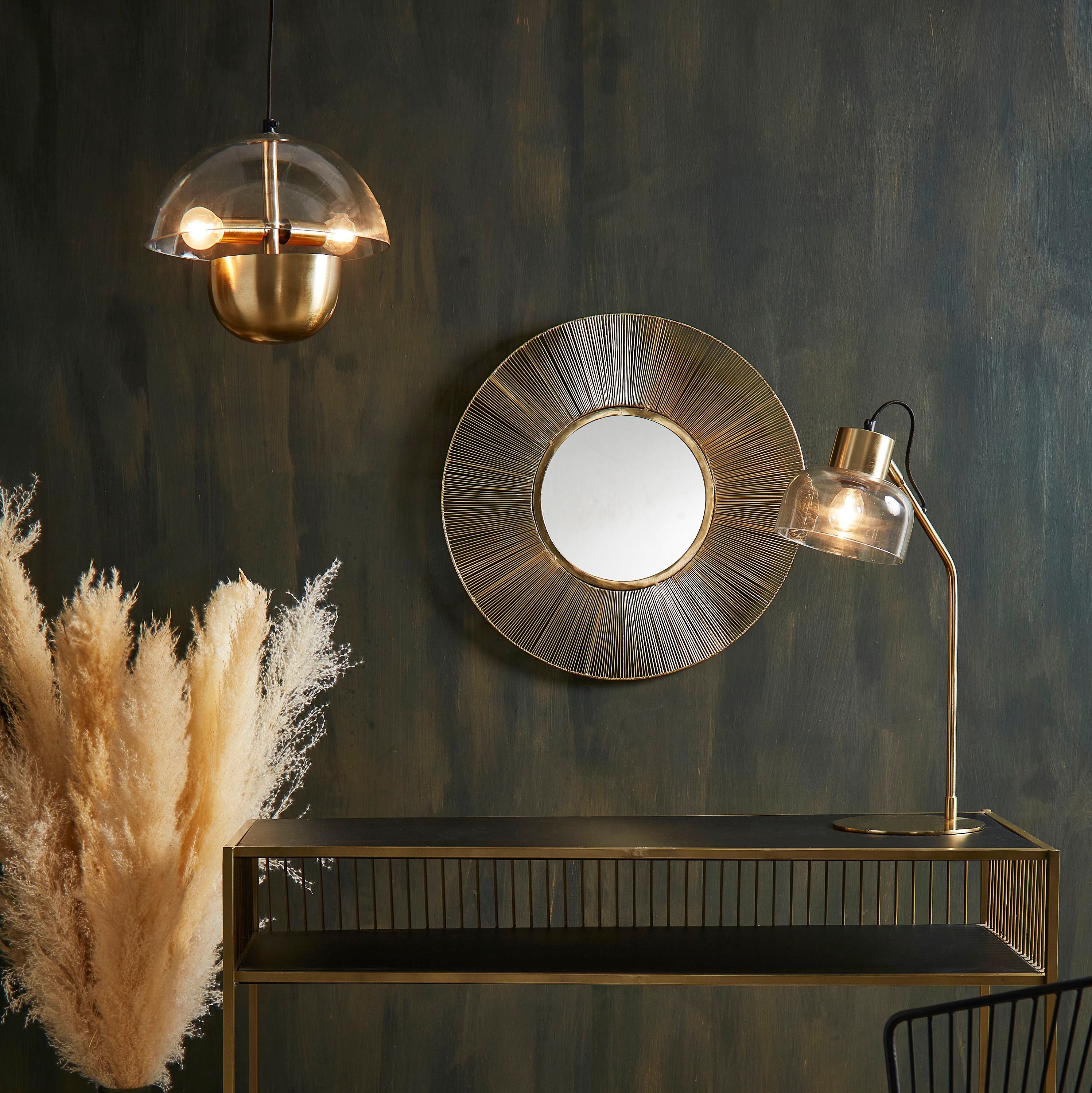 A styled interior scene featuring a brass console table against a dark textured wall, with a round sunburst wall mirror above it, a hanging glass pendant light, a floor lamp, a black wire chair with a patterned cushion, pampas grass in a vase, and a rug on the floor.